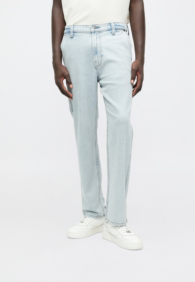 Jeans de mezclilla azul claro con un diseño de pierna recta, que cuentan con bolsillos delanteros y un cierre de botón. Se llevan con zapatillas blancas.