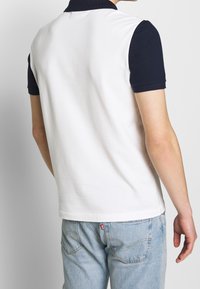 Polo à corps blanc et col bleu marine. Présente des manches courtes et un tissu texturé. Porté avec un jean en denim bleu clair.
