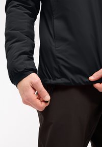 Zwarte buitenjas met een gestructureerde afwerking, voorzien van een trekkoord aan de onderkant. De hand past het koord bij de taille aan.