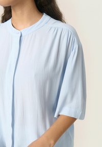 Camicia azzurro chiaro, con bottoni, colletto rotondo, maniche corte e tessuto testurizzato. Pieghe morbide evidenti lungo le spalle.