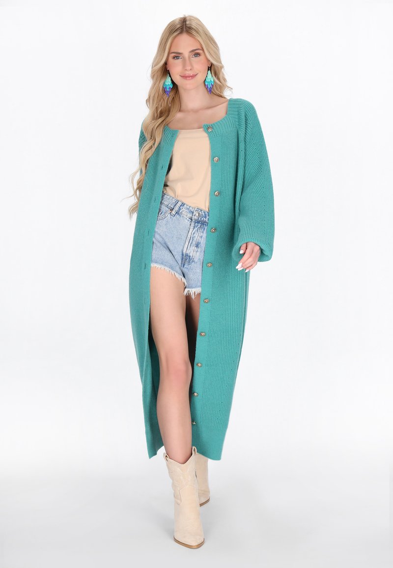 Cardigan lungo turchese con texture a coste, caratterizzato da grandi bottoni dorati, indossato sopra un top beige e pantaloni di denim, abbinato a stivali beige.