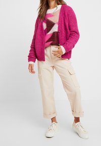 Cardigan a maglia fucsia con vestibilità ampia e superficie texture, indossato sopra un maglione multicolore a fantasia, abbinato a pantaloni cargo beige e sneaker.