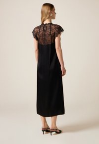 Zwarte jurk met een doorschijnend kanten inzetstuk en korte mouwen, met een striksluiting aan de achterkant en een lange, vloeiende silhouet.