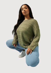 Olivgrön stickad tröja med ett öppet mönster, oversized passform och långa ärmar, tillsammans med ljusblå jeans och vita sneakers.