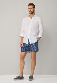 Weißes Leinenhemd, marineblaue Shorts mit weißem Herzmuster und blaue Espadrilles. Das Model steht mit einer Hand in der Tasche vor einem neutralen Hintergrund.