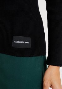 Pull noir côtelé avec une étiquette Calvin Klein Jeans sur le côté inférieur, porté avec un pantalon vert et une main droite visible sur un fond blanc.