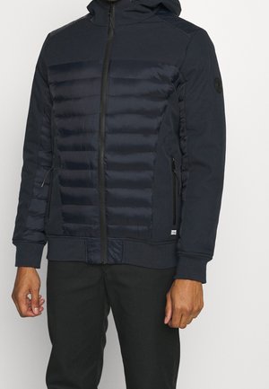 Mężczyzna ubrany w granatową pikowaną kurtkę z kieszeniami na zamek, ściągaczami na rękawach i kapturem, w połączeniu z czarnymi spodniami na jednolitym tle.