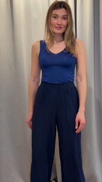 Mujer con pelo rubio hasta los hombros, vestida con una camiseta azul sin mangas y pantalones oscuros de pierna ancha, de pie frente a una cortina lisa de color claro.