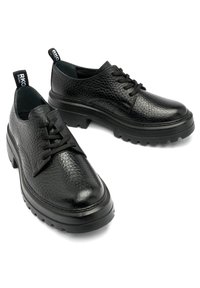 Chaussures en cuir texturé noir à lacets avec des semelles en caoutchouc épaisses et des tirettes de marque sur les talons, sur un fond blanc.