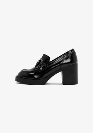 Zapato de tacón de charol negro con puntera redondeada, adornado con una hebilla decorativa y un tacón cuadrado texturizado.