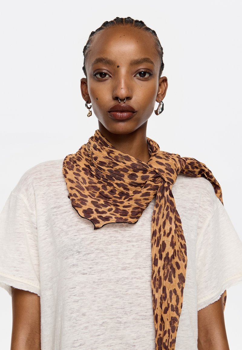 Leopard print scarf in shades of brown and tan, lightweight fabric with a wavy edge, knotted around the neck. Worn over a white top.