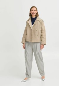 Veste beige à double boutonnage avec deux grands boutons, associée à un pantalon plissé gris clair et des escarpins argentés à bout pointu.