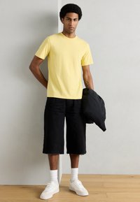 Κίτρινη βαμβακερή μπλούζα, μαύρα σορτς μέχρι το γόνατο, λευκά αθλητικά παπούτσια και κάλτσες μέσης. Η μπλούζα είναι απλή· τα σορτς έχουν ξεφτισμένο τελείωμα.