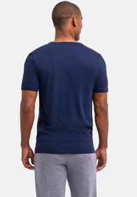 Mężczyzna w granatowej koszulce z krótkim rękawem i jasnoszarych dresowych spodniach, stoi tyłem do widza na jednolitym białym tle.