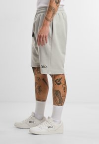 Lys grå bomullsshorts med elastisk midje, sidelommer og sort logodetalj, parret med hvite sneakers og hvite sokker.