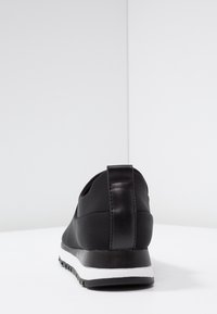 Chaussure de sport noire avec une tige synthétique lisse, un bout arrondi, une semelle intermédiaire blanche et une semelle extérieure en caoutchouc noir. Dispose d'une languette au niveau du talon texturée.