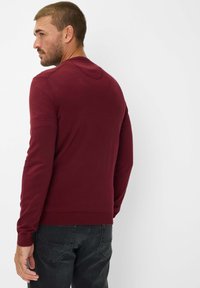 Bordeauxrotes langärmeliges Sweatshirt mit Rundhalsausschnitt, gerippten Bündchen und Saum. Glatte Textur, taillierte Passform. Kombiniert mit dunkelgrauen Jeans.