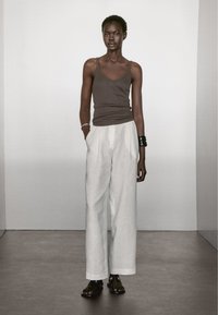 Bruine mouwloze top, loszittende witte linnen broek en geprinte sandalen. Accessoires zijn een zilveren armband en een zwarte armcuff.
