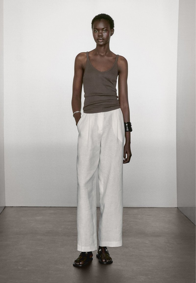 Bruine mouwloze top, loszittende witte linnen broek en geprinte sandalen. Accessoires zijn een zilveren armband en een zwarte armcuff.