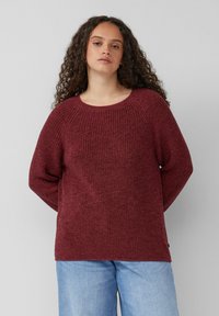 Burgunderfarbener Strickpullover mit Rundhalsausschnitt und Raglanärmeln, aus strukturiertem Stoff und in lockerer Passform. Kombiniert mit hellblauer Jeans.