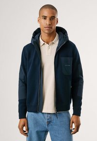 Hombre vestido con una sudadera con cremallera azul marino y parches acolchados en los codos, sobre una camiseta polo beige y jeans azules, de pie frente a un fondo claro y liso.