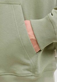 Vaalea vihreä huppari, jossa on ribbireunat hihansuissa ja helmassa, ja jossa on edessä tasku, jossa on näkyvät ompeluyksityiskohdat. Materiaali näyttää pehmeältä ja teksturoidulta.