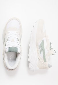 Sleek athletic sneakers in beige and white mesh with green suede accents. The design features a textured sole and lace-up closure.