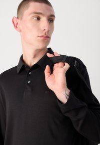 Fekete, hosszú ujjú polo póló gallérral, három gombbal és egy diszkrét logóval a mellkason. A modell egy fekete pántot tart a vállán.