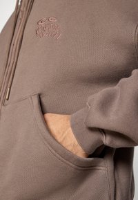 Camisola com capuz castanha com fecho de correr, costuras visíveis, bolso canguru e logótipo bordado no peito, feita de tecido texturizado.