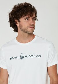 Bílá bavlněná trička s kulatým výstřihem, s výrazným tmavě šedým logem "SAIL RACING" přes hruď. Krátké rukávy, klasický střih.
