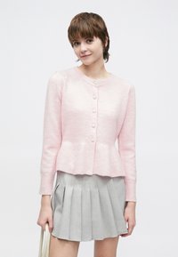 Różowy sweter na drutach z talią typu peplum, z okrągłym dekoltem i sześcioma ozdobnymi guzikami. Nosi się go na plisowaną szarą spódnicę.