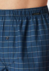 Boxers à carreaux bleus en tissu doux, avec une ceinture élastique, une fermeture à bouton et de fines rayures blanches et grises sur un fond bleu plus foncé.