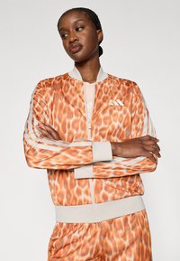 Sieviete ar bizelētu matu stilu valkā oranžu un bēša rakstu sporta tērpu, ar sakrustotām rokām, skatoties nedaudz uz sāniem pret vienkāršu fonu.