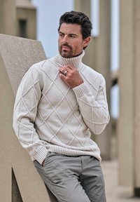 Hellbeige Rollkragenpullover mit einem Rautenmuster, gerippten Bündchen und Saum, kombiniert mit hellgrauen Hosen, vor einem neutralen Hintergrund.