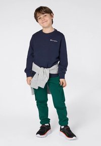 Sweatshirt azul-marinho com logotipo branco, sweatshirt cinzento amarrado à cintura, calças cargo verdes e sapatilhas pretas com sola branca e detalhes em laranja.