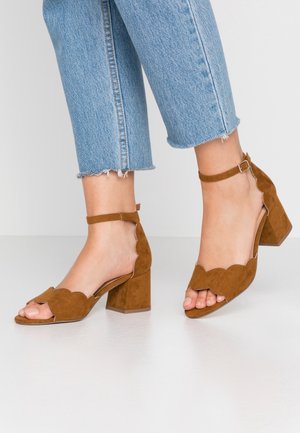 Bruine suède hoge hakken sandalen hebben een geschulpte ontwerp en een enkelbandje, met een chunky zool en open teen, gecombineerd met cropped denim.