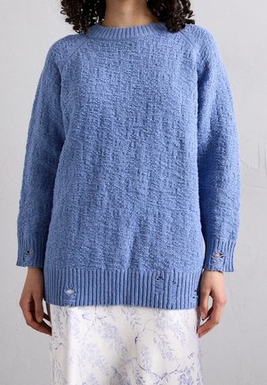 Femme portant un pull bleu clair texturé avec des trous usés et une jupe blanche avec un motif floral léger.