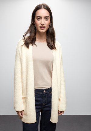 Krämfärgad, fluffig kofta med öppen front, mysig textur, i kombination med en beige topp och mörk denimjeans. Har två framfickor.
