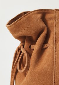 Sac en suede beige avec une texture douce, doté d'une fermeture à cordon, d'une couture soignée et de détails plissés à l'ouverture supérieure.