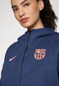 Navy zip-up hoodie with orange Nike logo and Barcelona emblem on the chest. Features a soft fabric and a high, curved collar.