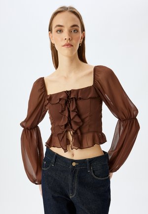 Bruine ruches blouse met een vierkante halslijn, lange doorschijnende mouwen en een strikdetail aan de voorkant, gecombineerd met donkere jeans. Heeft een gestructureerde stof.