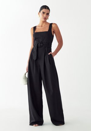 Naine seisab, kandes musta varrukateta kombekat laiade säärtega, vööga taljes, kuldsete rõngaskõrvarõngastega, hoides käes väikest valget käsikotit.