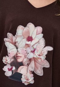 Brązowy sweter z dzianiny z aplikacją kwiatową w różowe i białe kwiaty, motylami oraz nadrukowaną twarzą, wyśrodkowaną z tekstem "wszystko w sercu."