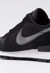 Black Nike sneaker featuring a glittery silver swoosh logo, suede and mesh upper, and white cushioned sole with black tread.