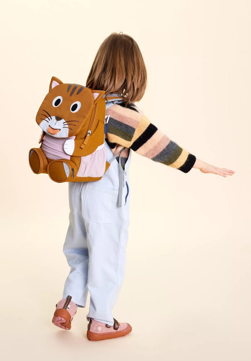 Kind mit schulterlangem Haar, das einen gestreiften Pullover und helle Latzhosen trägt, einen braunen katzenförmigen Rucksack tragend und einen Arm seitlich ausstreckend.