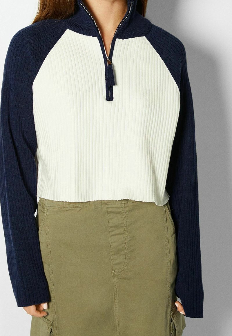 Kvartsdels zip-tröja i kräm och marinblå med ribbad textur; har lång ärm och cropped design. Kombinerad med en olivgrön kjol.