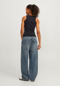 Mørk strikket ermeløs topp kombinert med løse blå denimjeans, med høy midje og store baklommer, iført hvite og svarte joggesko.