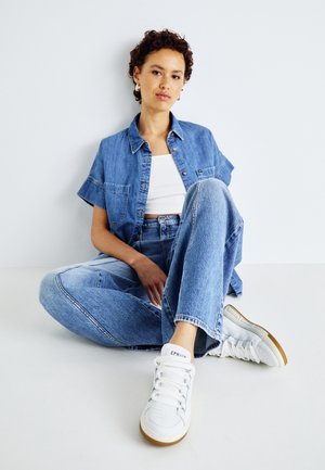 Mujer joven sentada en el suelo con una camisa de manga corta de denim, vaqueros de talle alto, top corto blanco y zapatillas blancas con suela de goma.