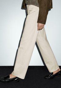 Beige trousers made of smooth fabric with a straight cut, paired with black loafers. The model walks against a minimalist background.