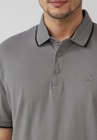 Grijze poloshirt met een gestructureerde stof. Heeft een kraag met zwarte accenten en een twee-knoops sluiting. Subtiel logo op de borst.